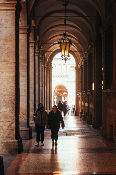 Two figures walking under Bologna's famous vaulted porticoes, warm golden light at the end of the arcade