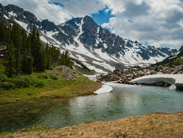 A clear mountain stream winds through a rocky alpine meadow with remnant snowfields, dramatic Beartooth peaks rising sharply behind under a partly cloudy sky