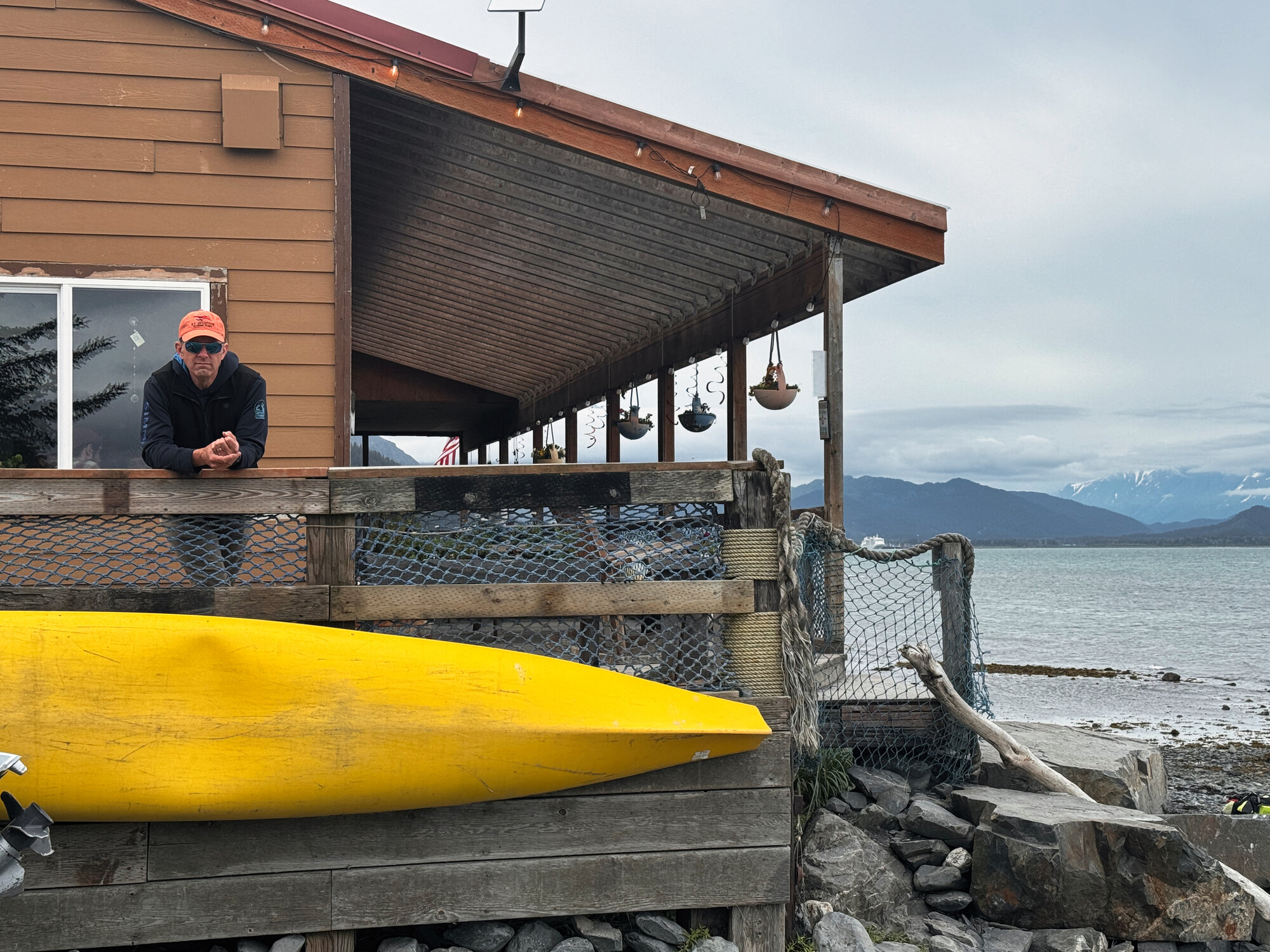 Nick leaning on the deck railing of Miller's Landing lodge in Alaska, yellow kayak in the foreground, mountains and inlet stretching behind