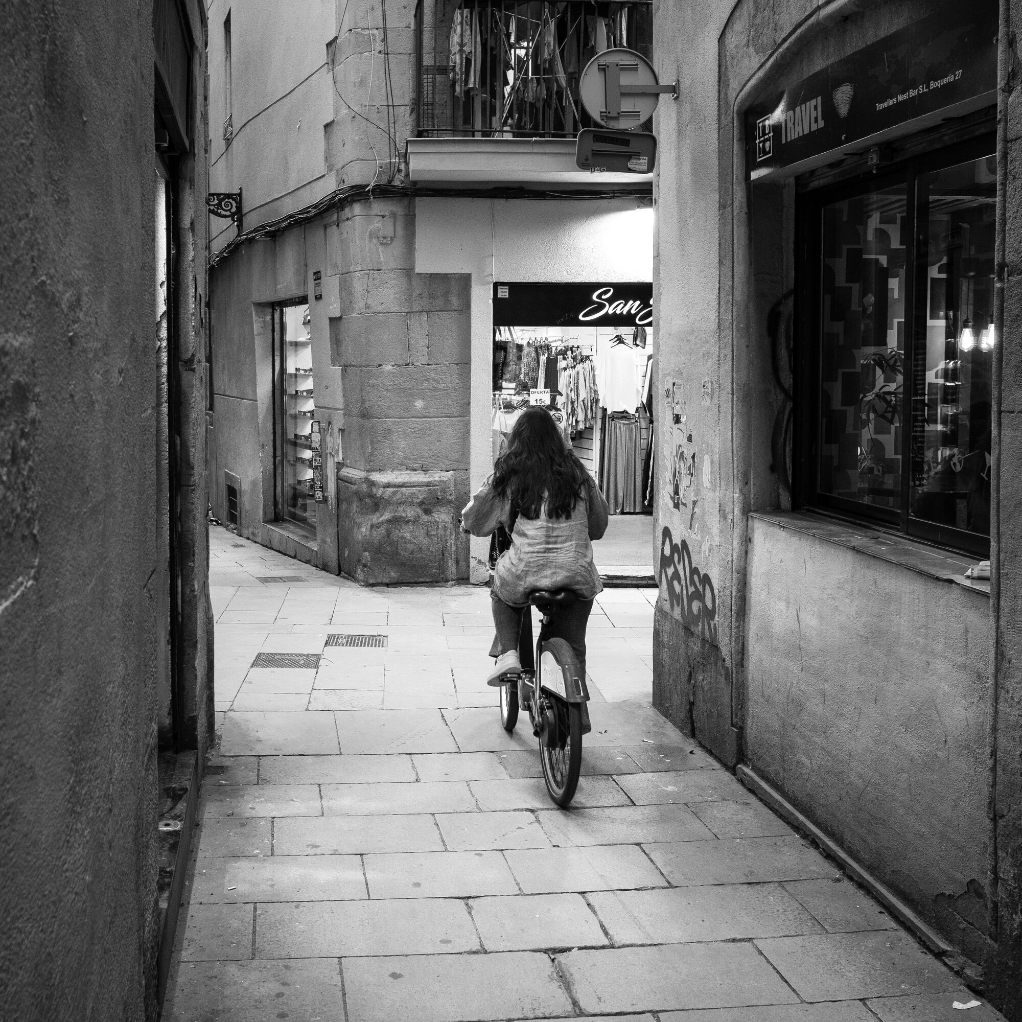 Woman on a bike riding away through a narrow Gothic Quarter alley flanked by stone walls and shopfronts, heading toward a lit storefront at the end of the lane