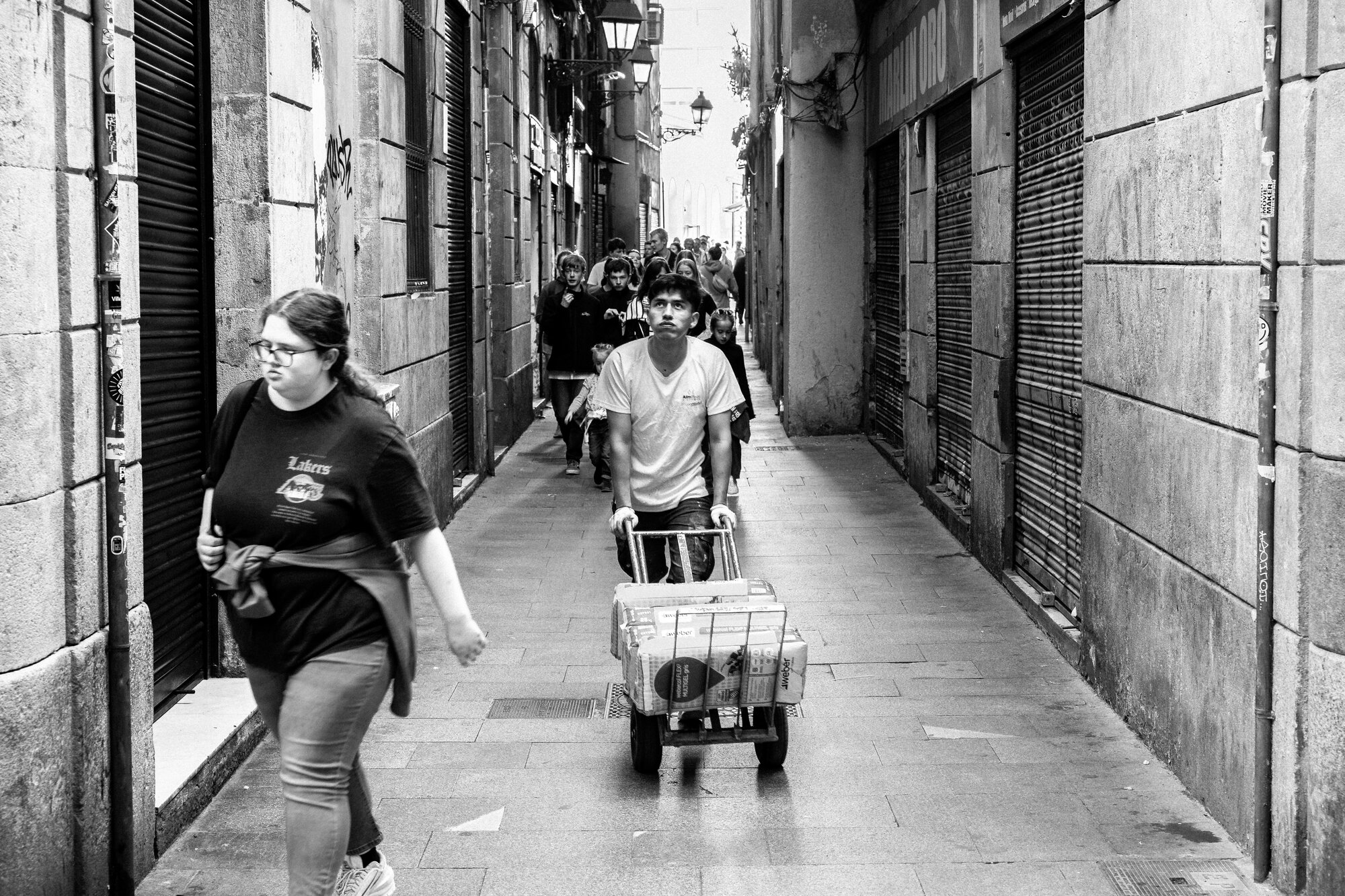 Gothic Quarter narrow alley with a worker pushing a delivery cart past medieval stone walls, overhead lamp casting pools of light