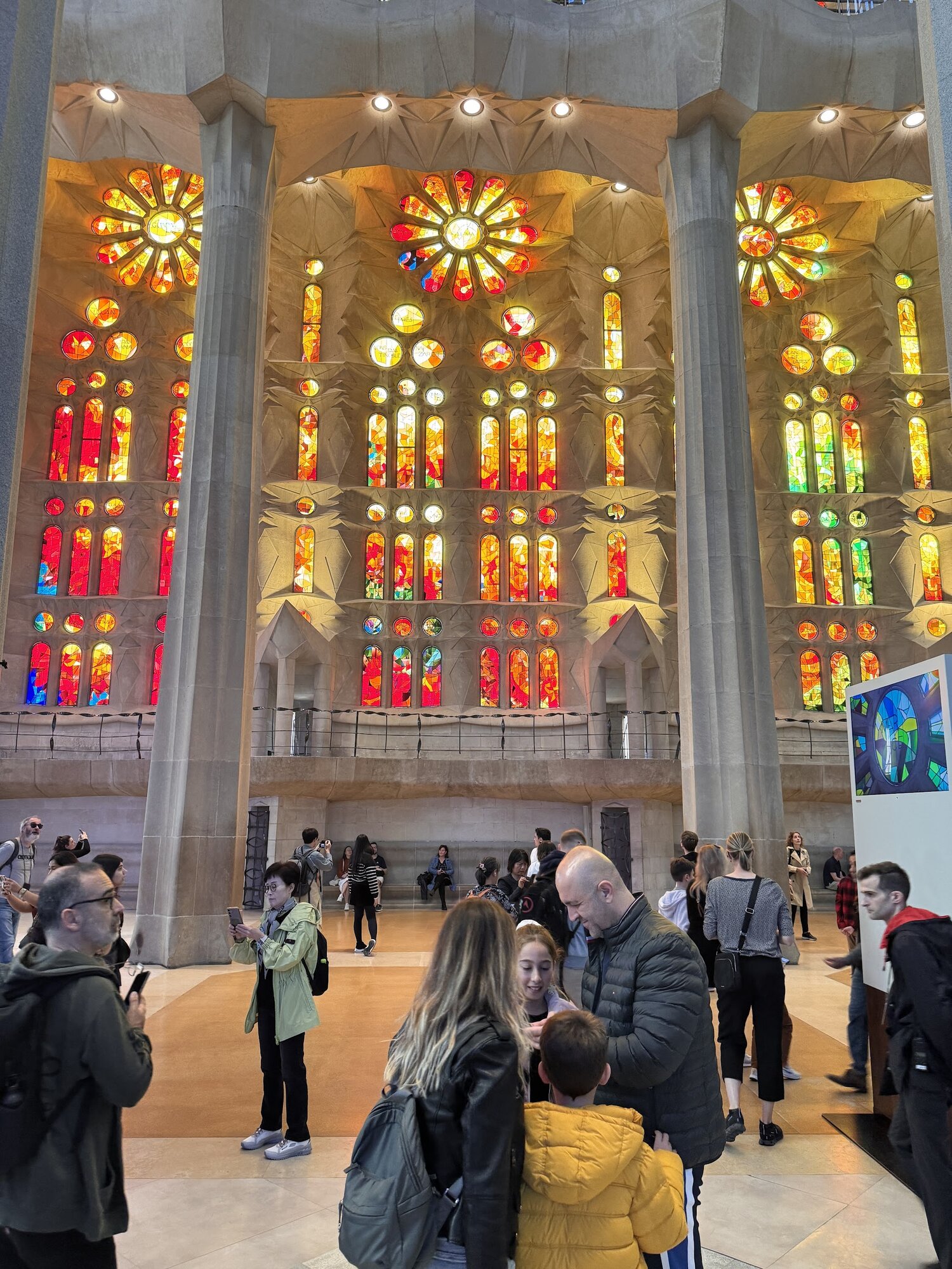 Interior of Sagrada Família with warm red and orange stained glass windows glowing above soaring stone columns