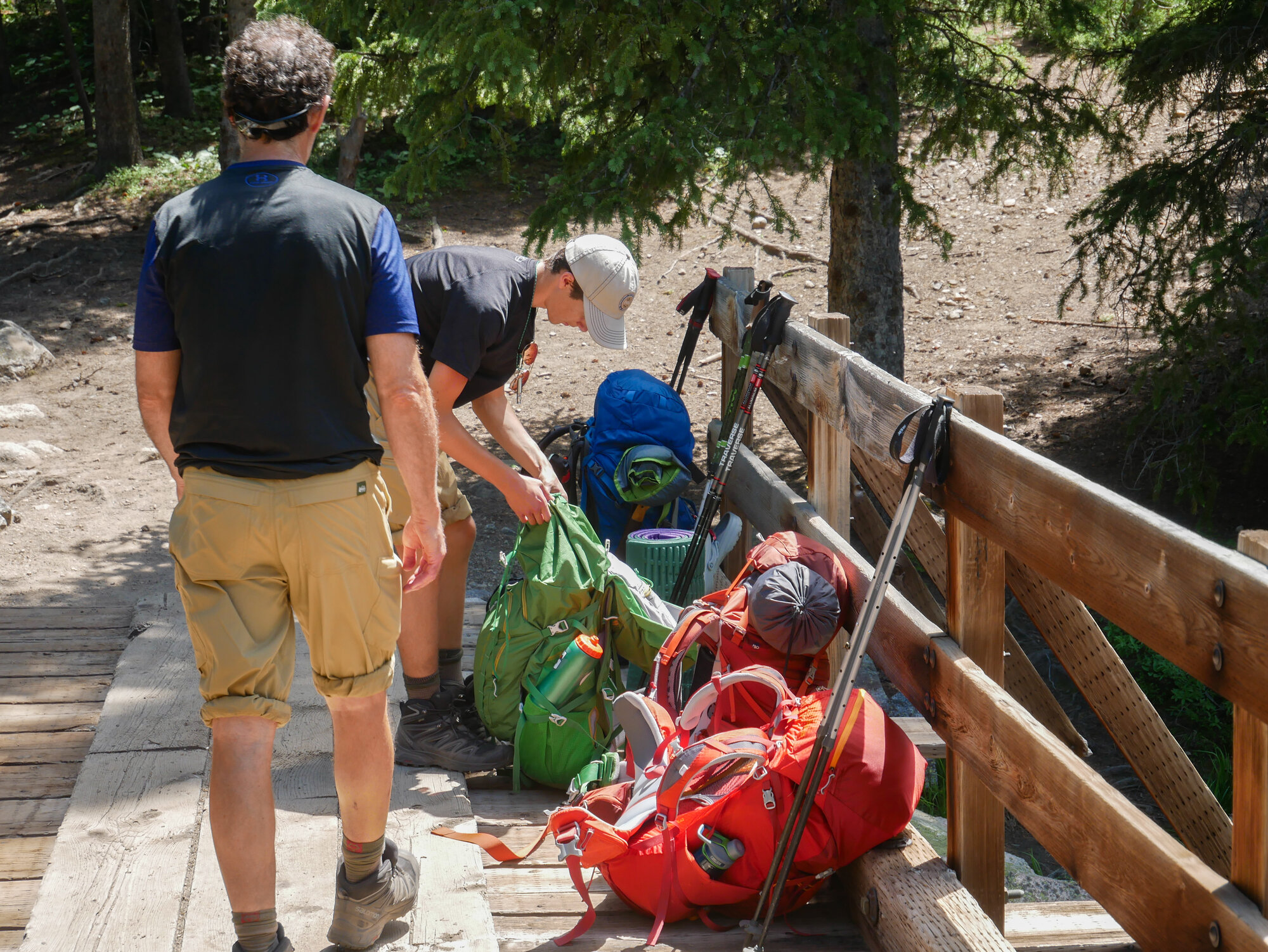 Two hikers on a wooden trailhead bridge with enormous overstuffed packs piled beside them — the physical reality of carrying too much into the backcountry