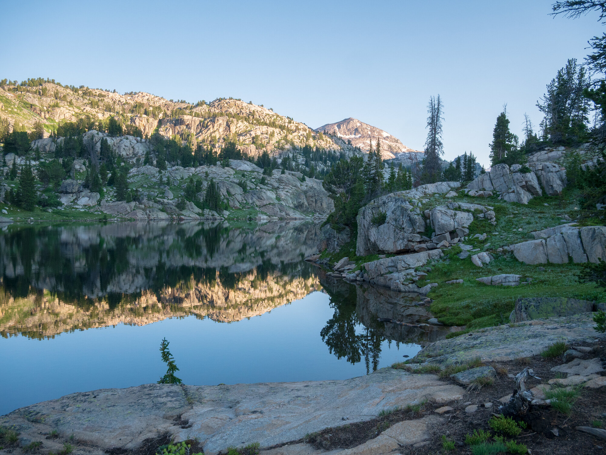 Becker Lake at first light, granite peaks and pine ridgeline reflected in still water on the Beartooth Plateau