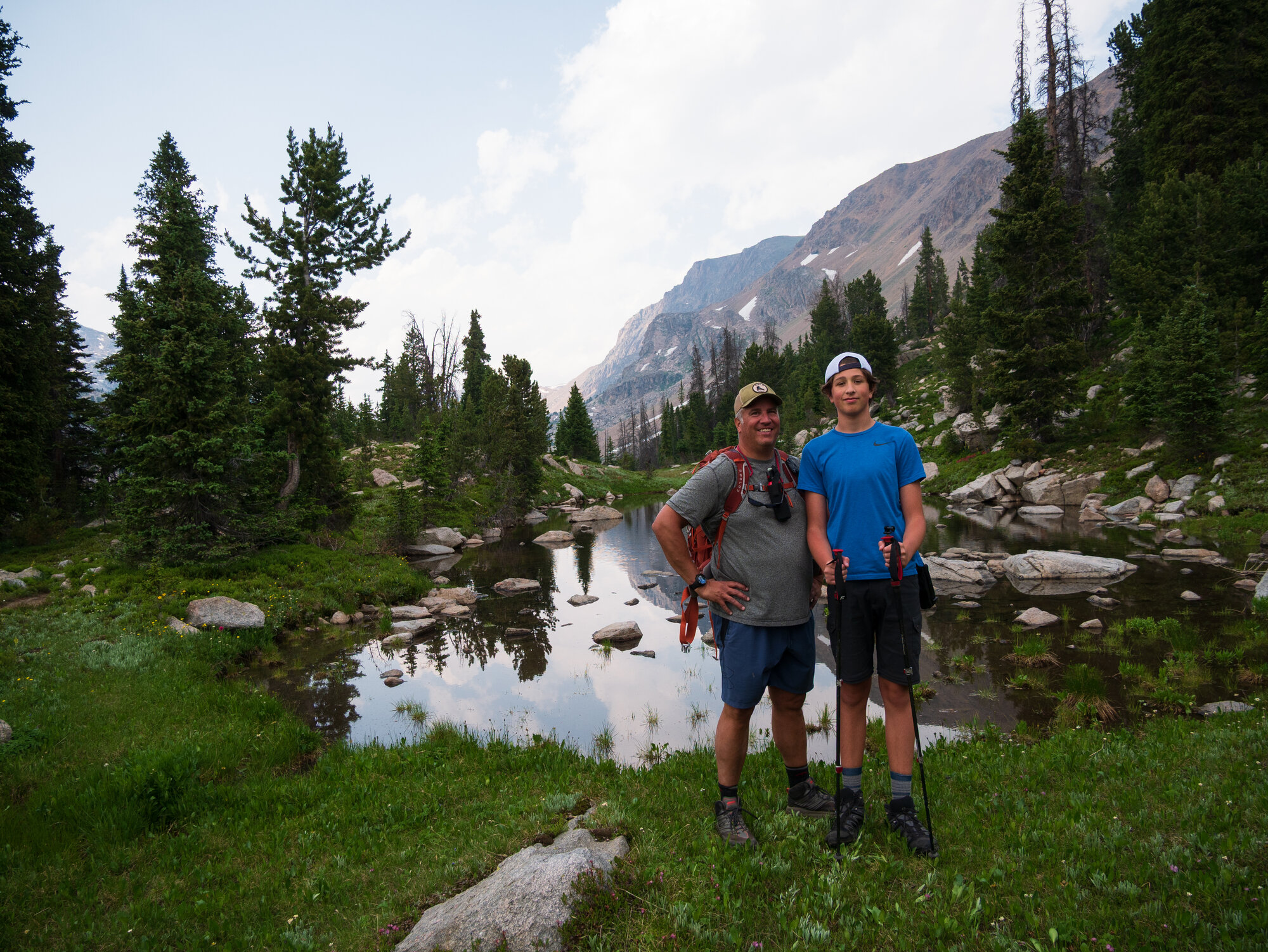 Nick and Niko standing together in front of a small tarn reflecting the mountains and trees behind them at dusk