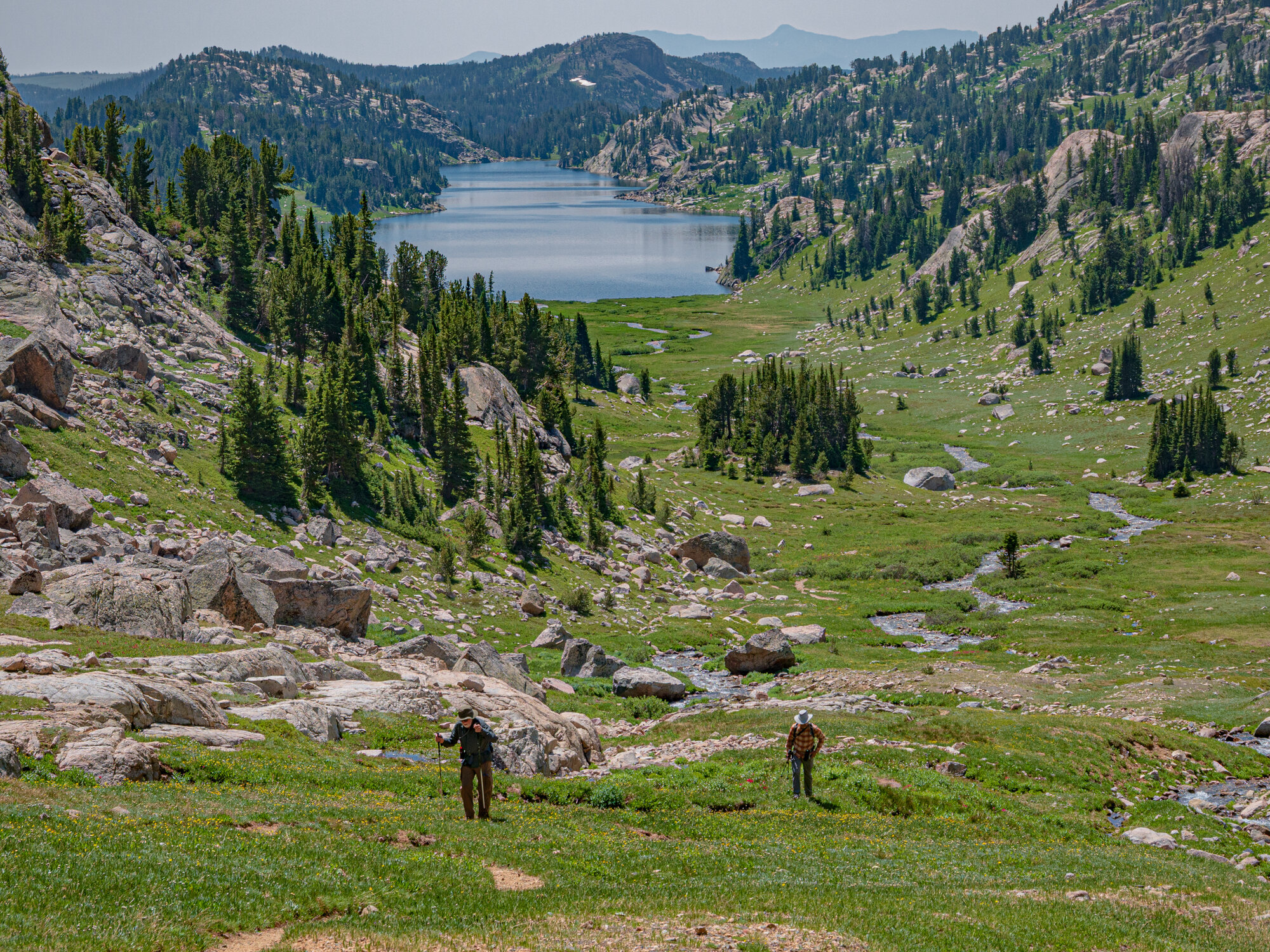 Two small hikers descend a wide green alpine valley toward Becker Lake, clearly visible as a blue oval in the distance below