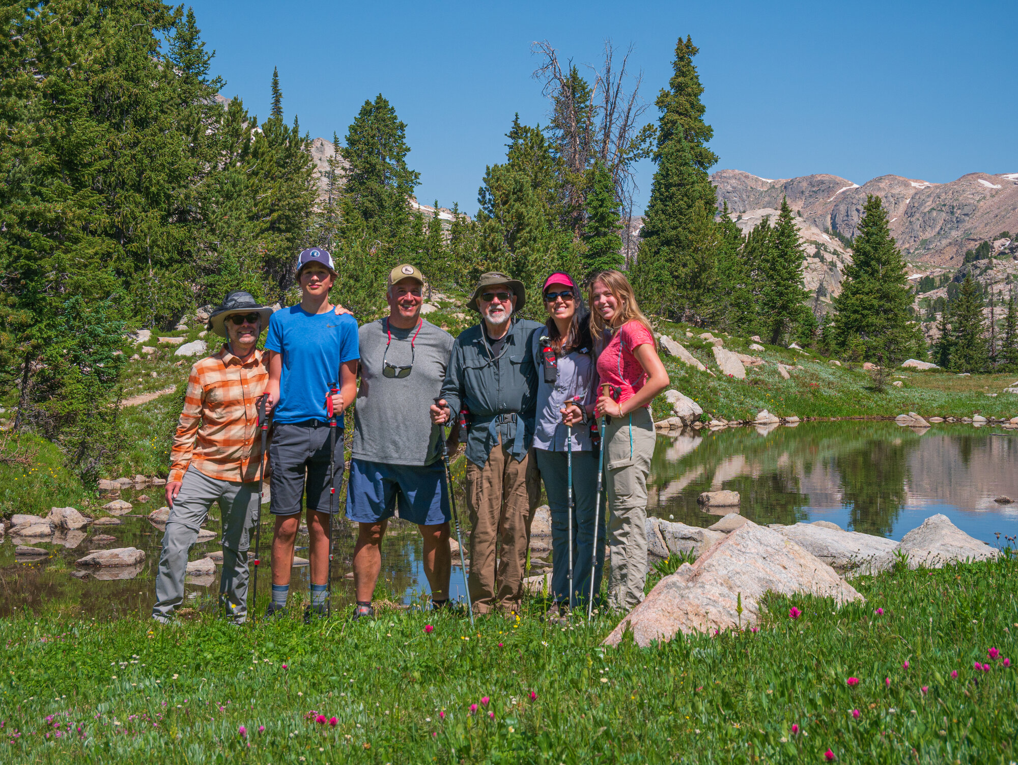 A group of six hikers smiling together in an alpine meadow with a small tarn, wildflowers and mountain peaks behind them