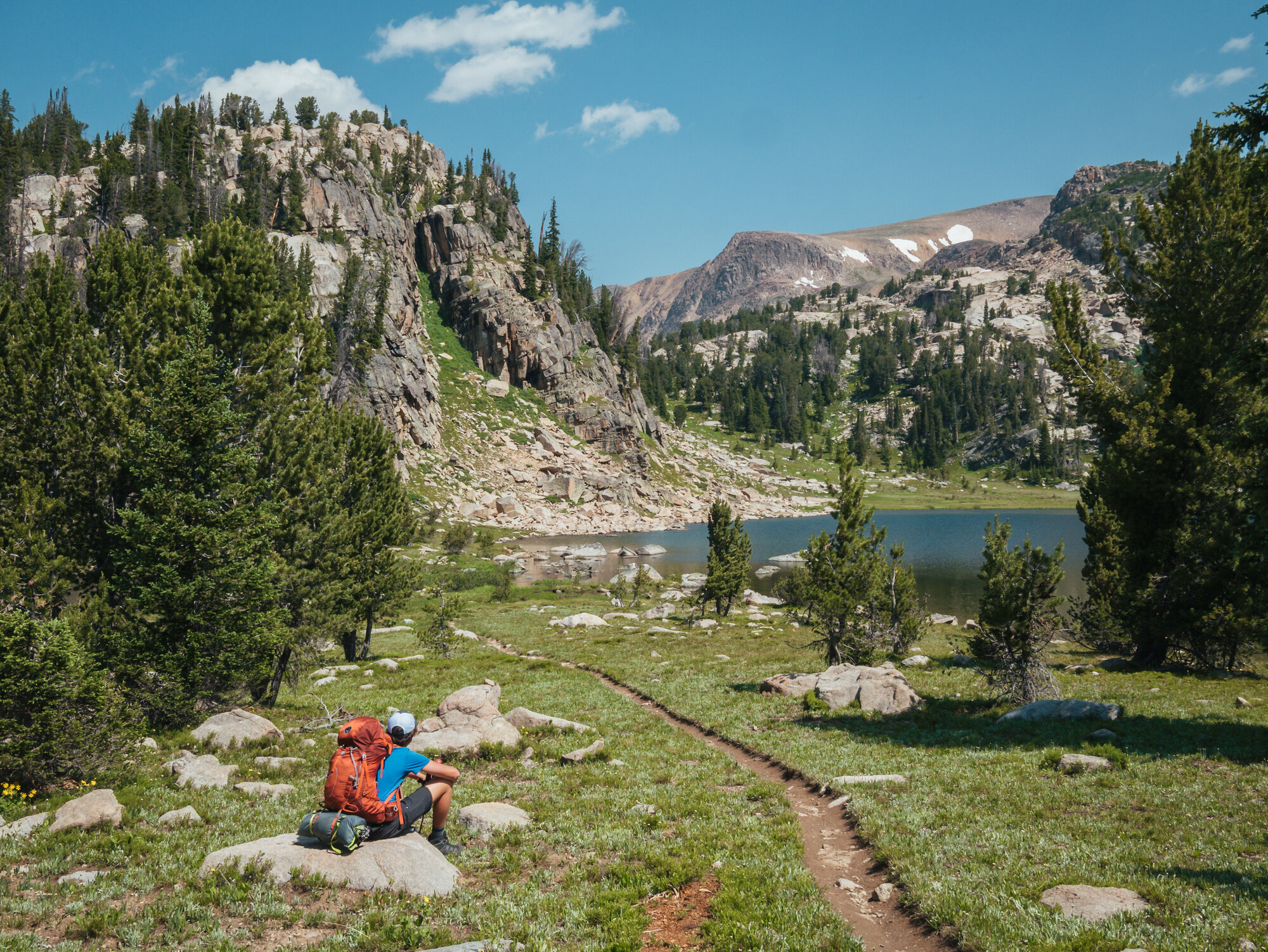 A hiker with a full red backpack rests on a boulder looking out at a pristine alpine lake framed by granite cliffs and snow-dusted peaks