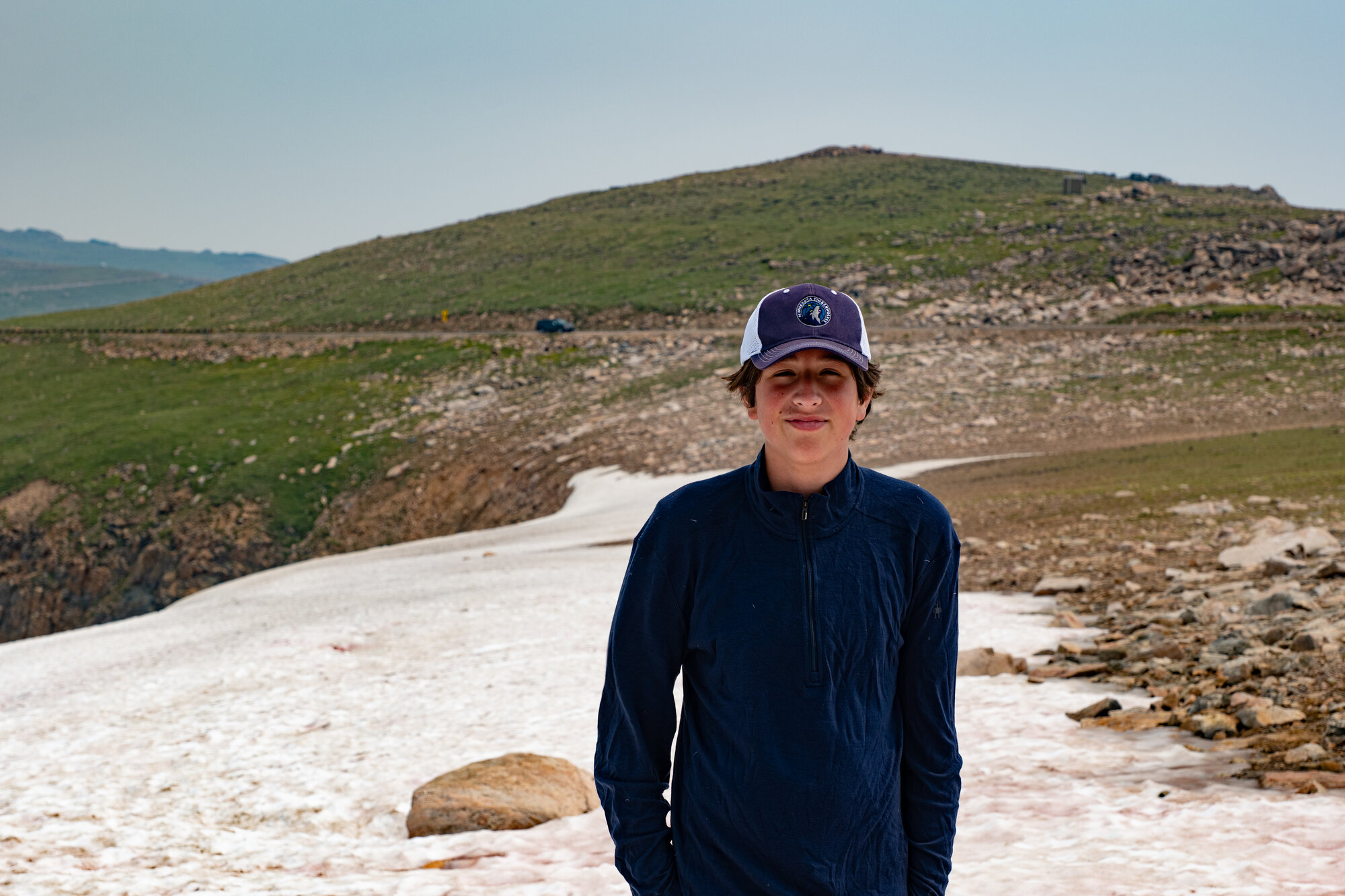 Niko standing on a snowfield on the open Beartooth Plateau, a rounded green peak and hazy smoky sky behind him