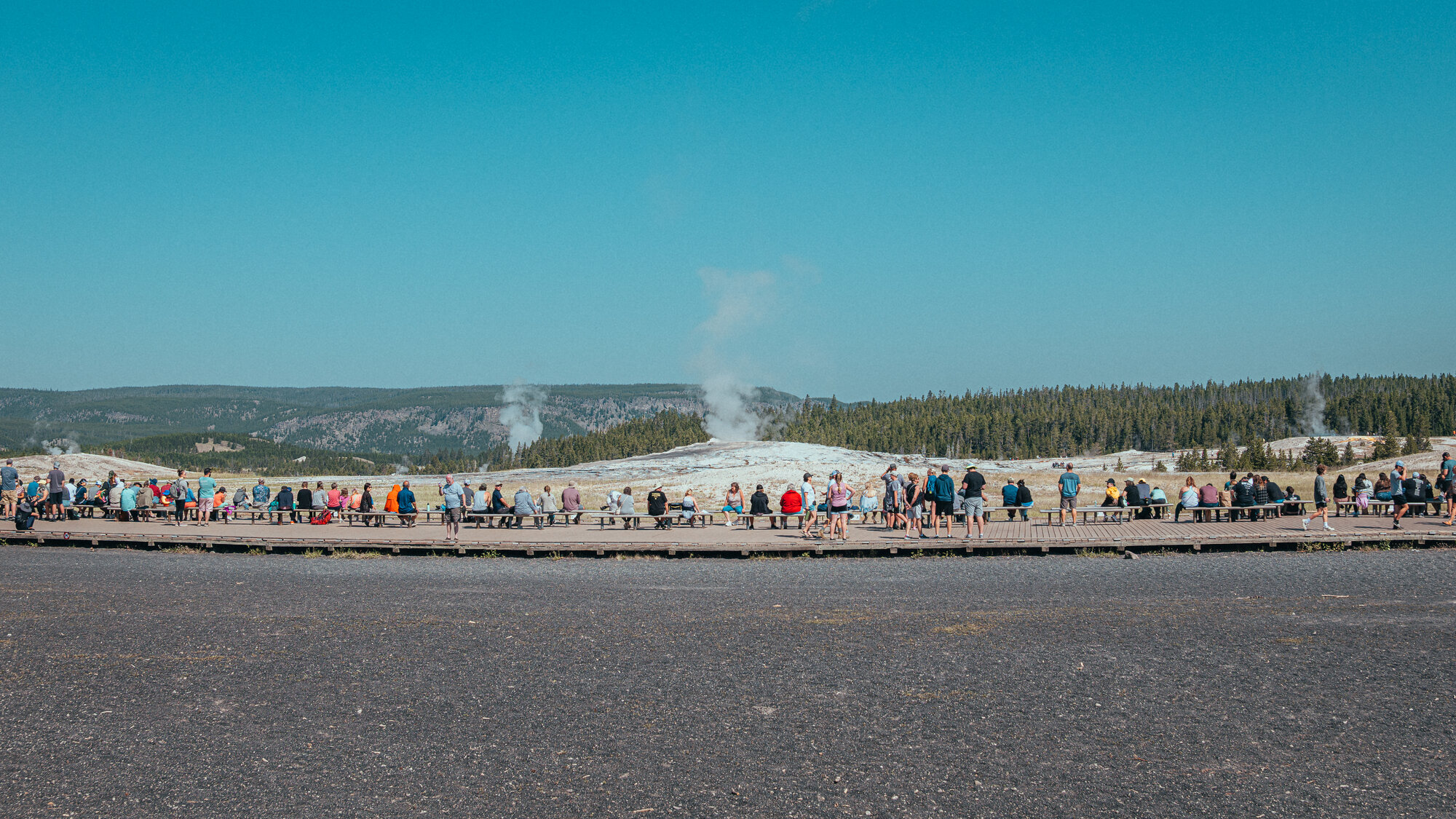 Wide panoramic view of the Old Faithful viewing area packed with visitors, the geyser beginning to erupt in the center distance