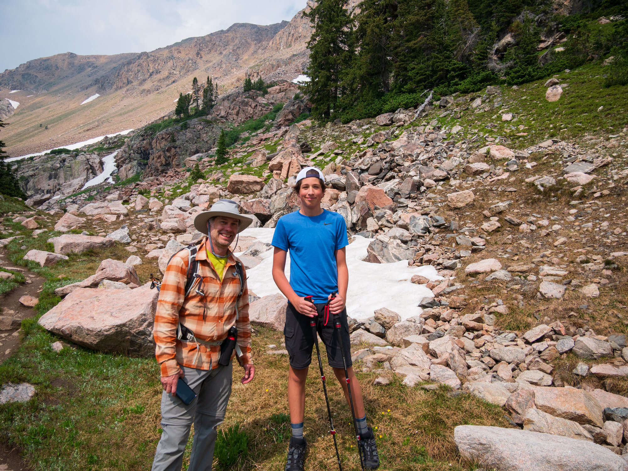 Nick and Niko standing on a snowfield near Albino Lake, a mountain waterfall and rocky peak rising dramatically behind them