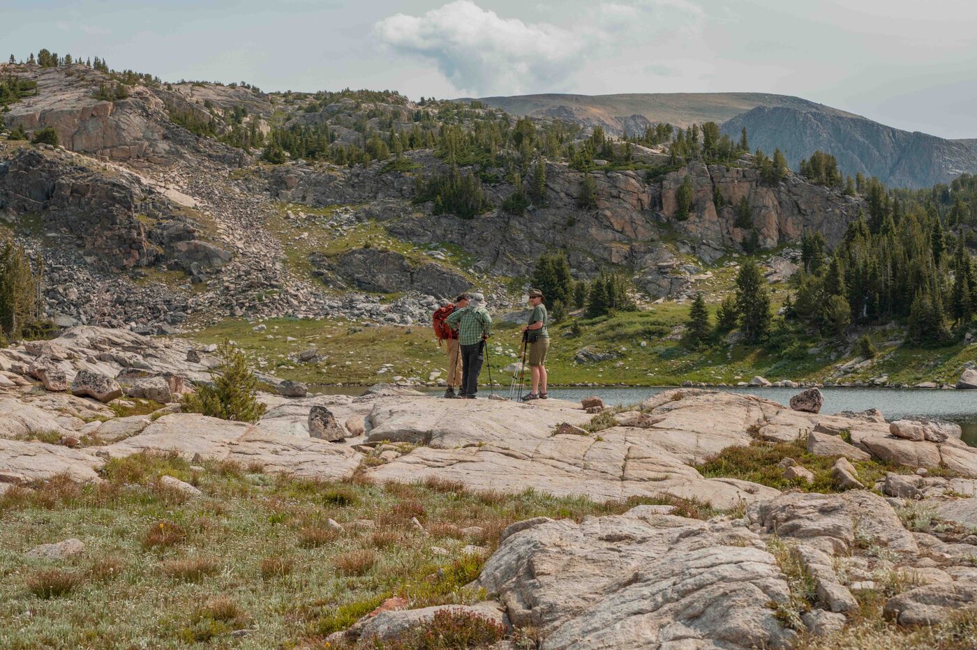 Two hikers standing on wide granite slabs near a calm alpine lake on the Beartooth Plateau, consulting a map or phone, rocky ridgeline and open tundra stretching behind them