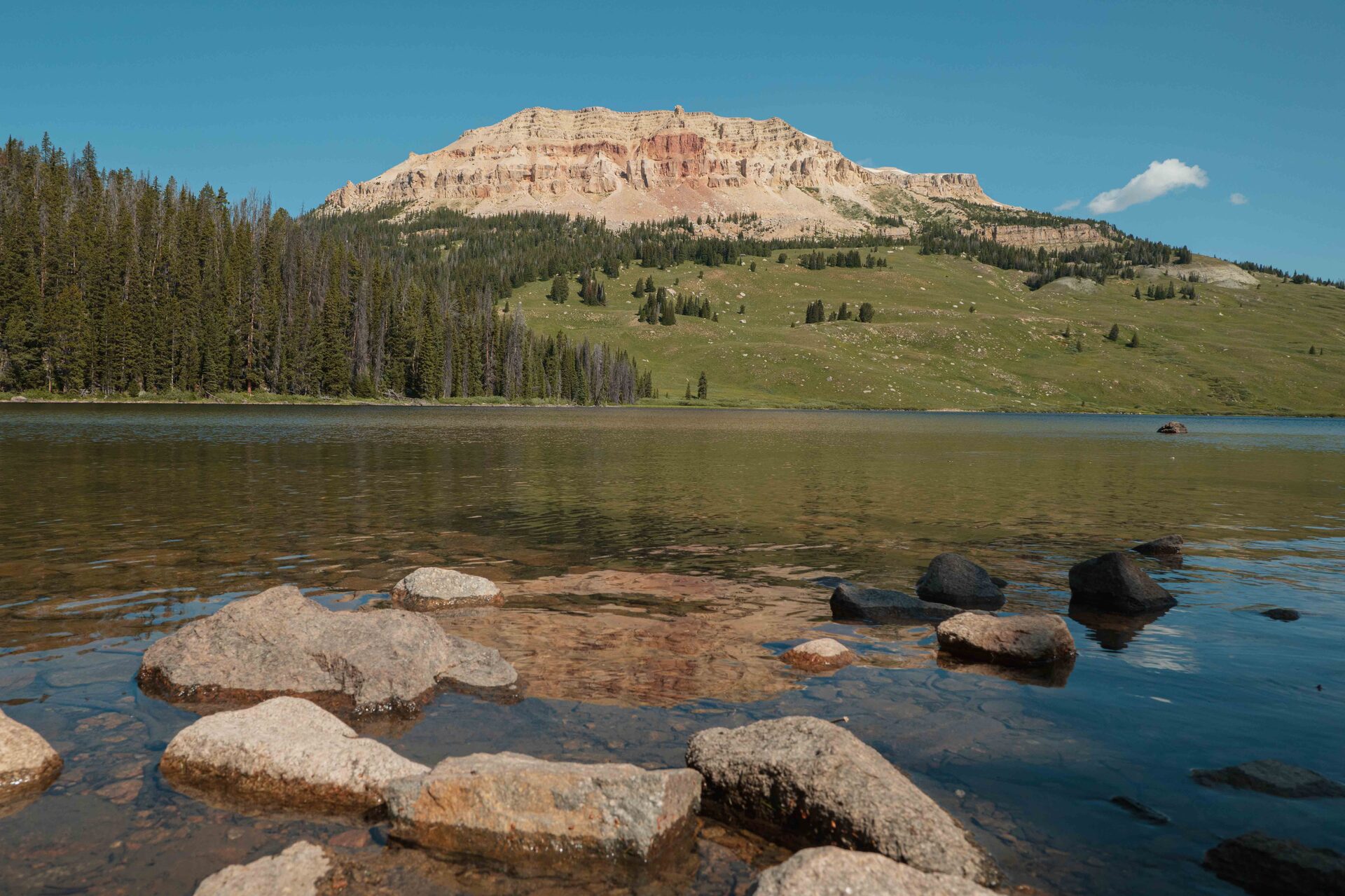 Beartooth Lake with rounded boulders in clear shallows and the dramatic layered red and tan cliffs of Beartooth Butte rising above the far shore