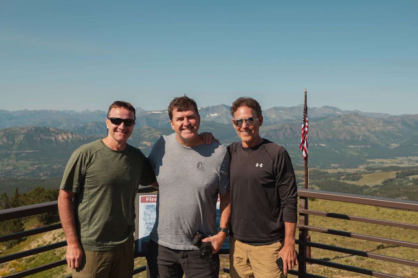 Three hikers posing together at the Clay Butte Fire Lookout observation deck, the Beartooth Plateau and distant mountain ranges stretching to the horizon behind them under a clear blue sky