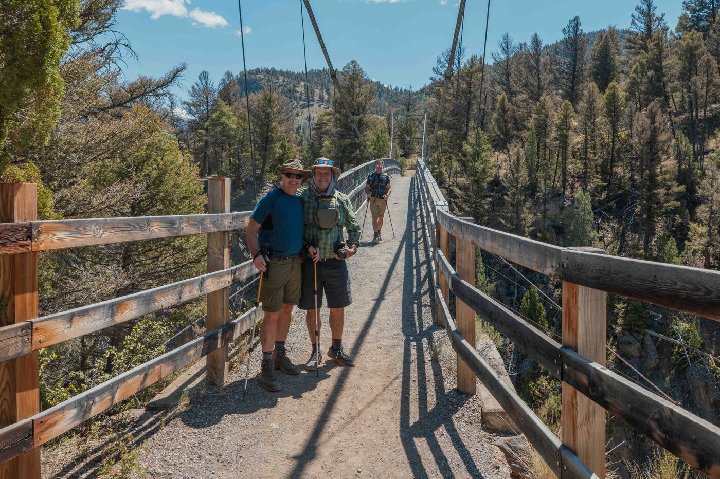 Two hikers posing on the Hellroaring suspension bridge over the Yellowstone River, wooden railing stretching behind them, pines and canyon walls rising on both sides