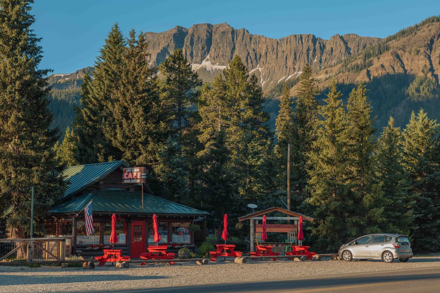 The Log Cabin Café in Cooke City at early morning, red picnic tables outside, dense pines and a rugged mountain peak rising sharply behind the small building under a deep blue sky