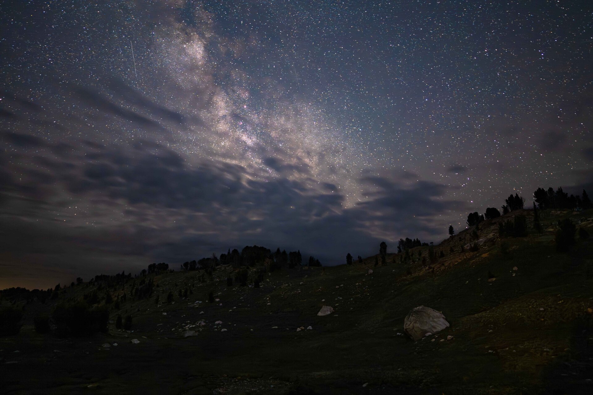 The Milky Way arcing brilliantly across the sky above the Beartooth Plateau, thin clouds parting around the galactic core, pine trees silhouetted on the dark ridgeline, a single boulder in the foreground