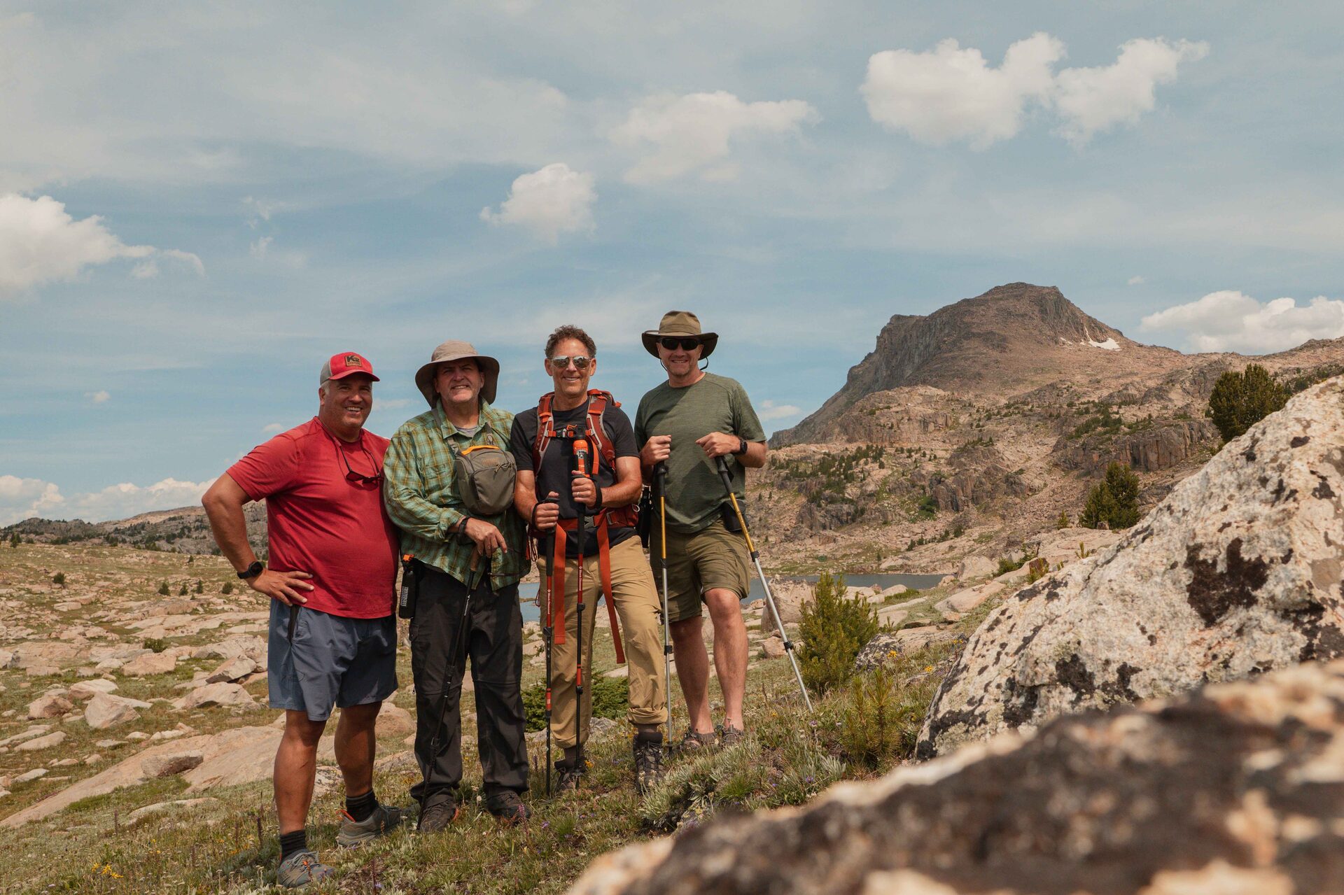 Four hikers posing together on the open Beartooth Plateau, an alpine lake visible in the valley below and a dramatic rocky peak rising behind them under a partly cloudy sky