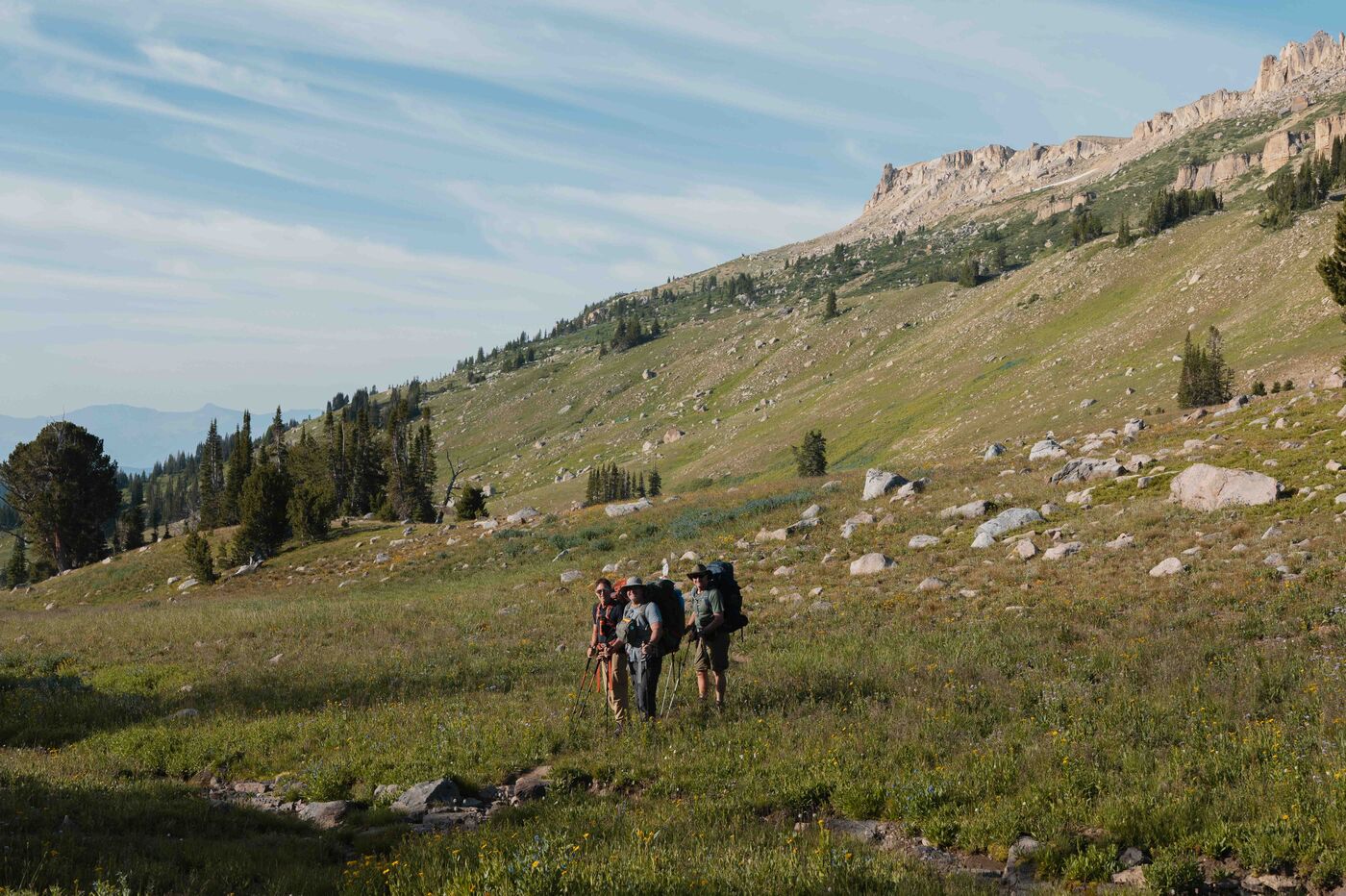 Three hikers with full backpacks walking through a wide open alpine meadow, a rocky ridgeline rising above and to the right, wildflowers dotting the grass in the foreground