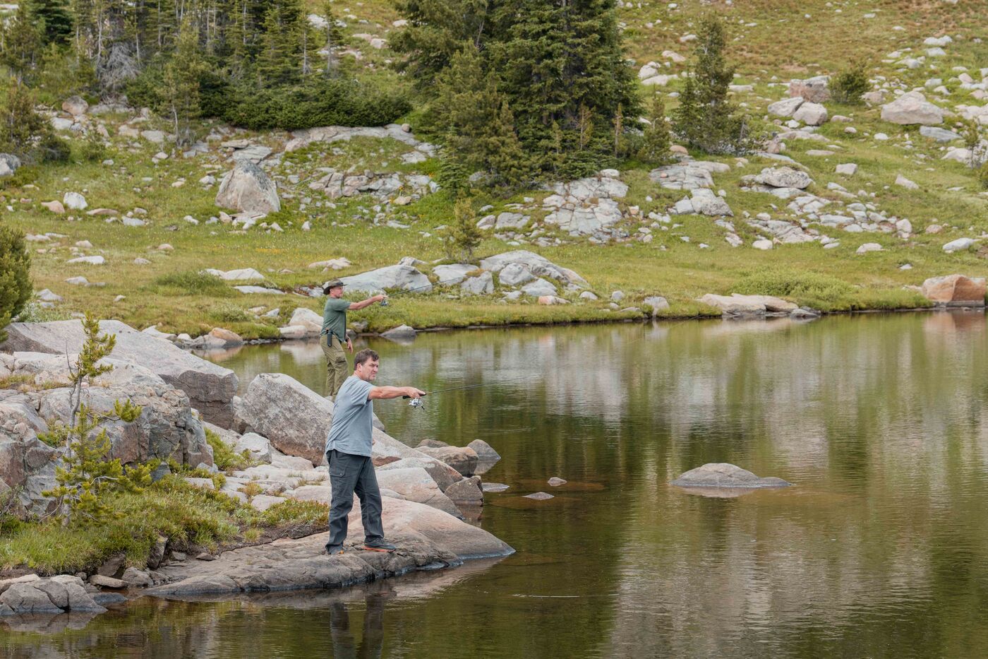 Two anglers casting from rocks along a calm, boulder-lined alpine lake on the Beartooth Plateau, green tundra and granite hillside stretching behind them