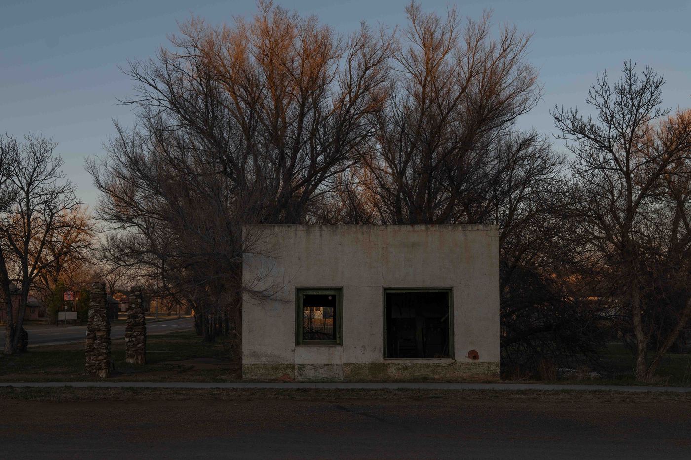 An abandoned concrete building at dusk on a quiet street, bare cottonwood trees glowing amber in the last light behind it