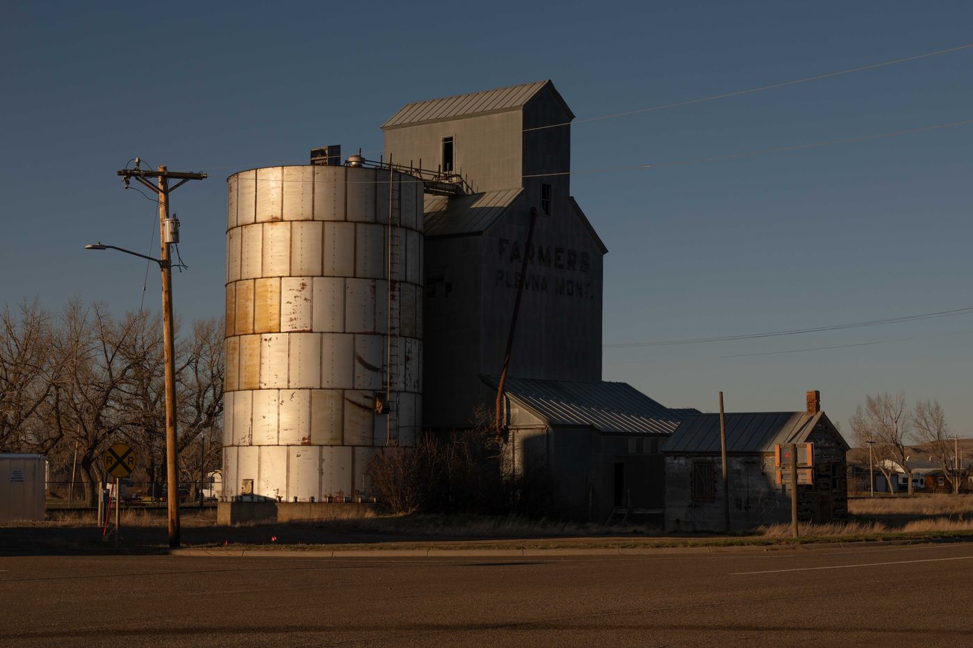 A weathered grain elevator and metal storage silo at golden hour in a small North Dakota town, warm light catching the metal