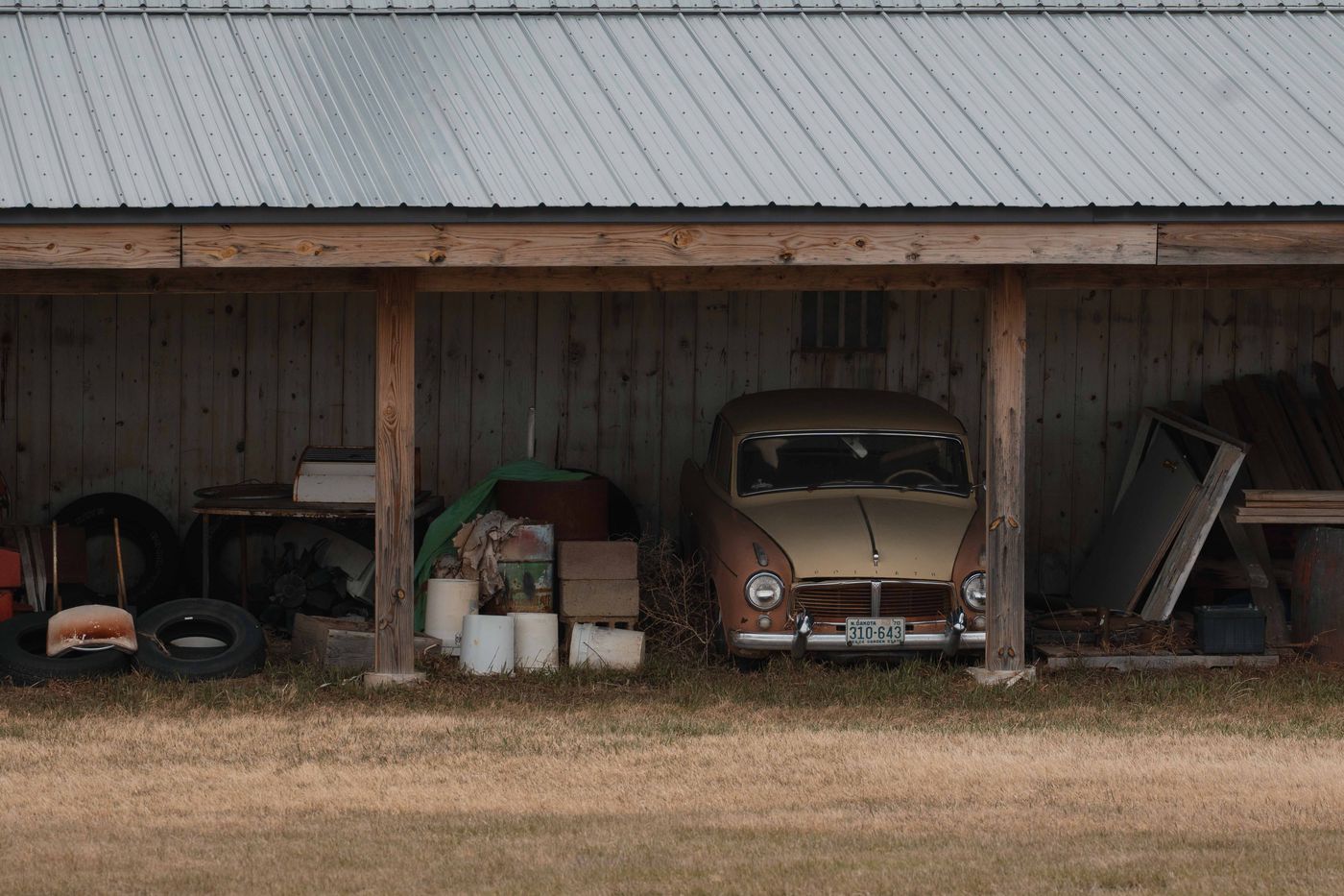 A vintage gold-colored car parked beneath a wooden farm shed with a corrugated metal roof, surrounded by old tires and farm debris