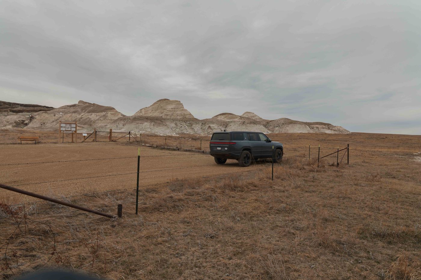A dark Rivian SUV parked on a dirt pullout at White Butte with vast flat North Dakota prairie stretching to the horizon behind it under a wide open sky