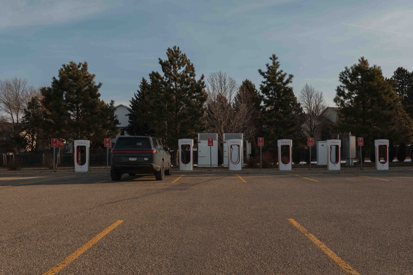A Rivian SUV pulling into a Tesla Supercharger bank in an empty parking lot at dusk, a row of white charging stalls illuminated against dark trees