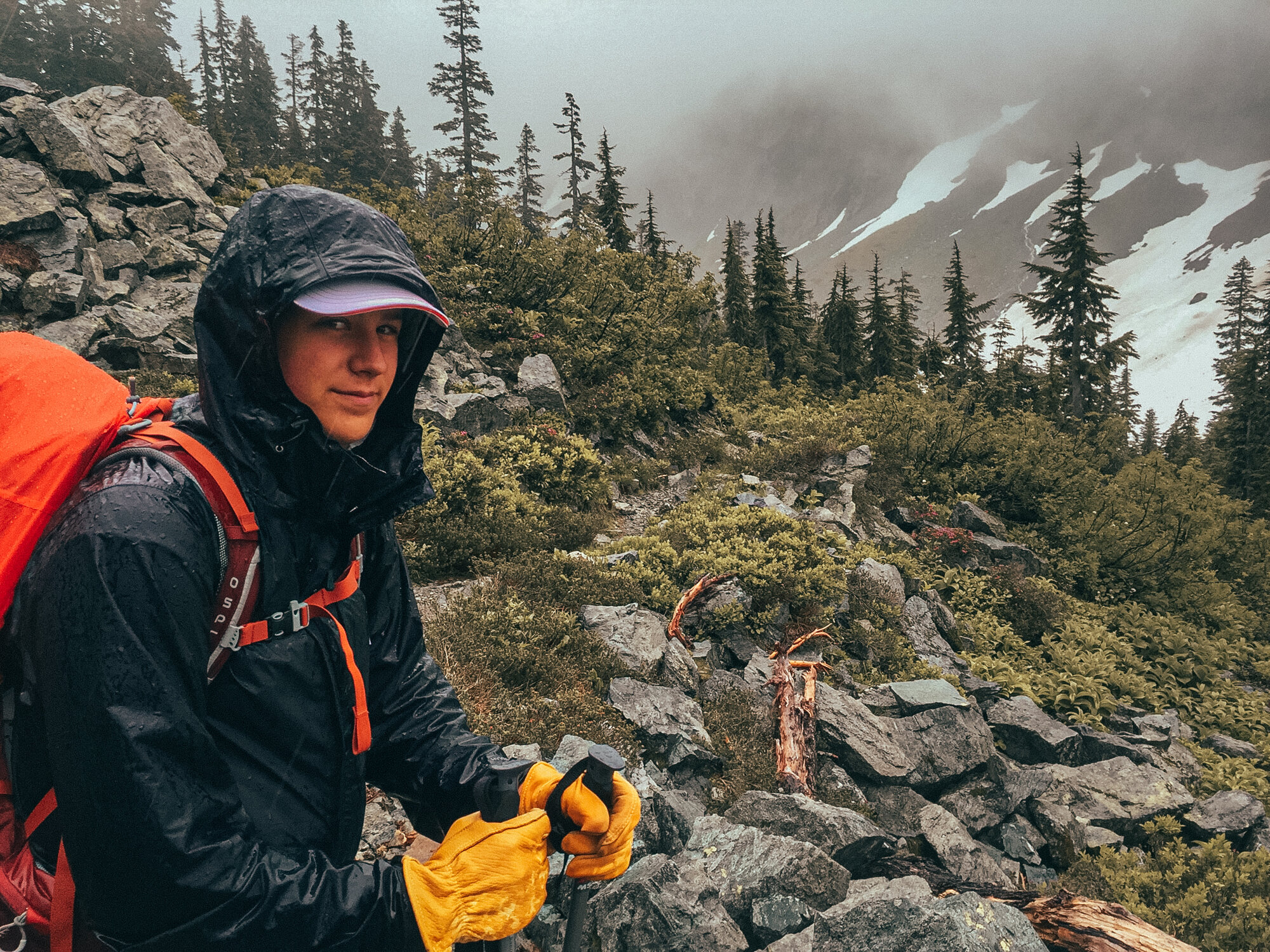 Hiker in rain gear with yellow gloves and a red pack pausing on a rocky alpine slope, snow and conifers in the mist behind them